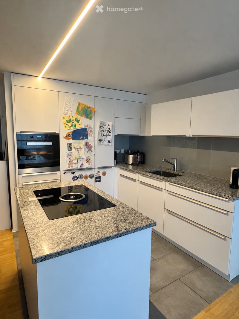Modern kitchen with white cabinets, granite countertops, an induction cooktop, a stainless steel oven, and a refrigerator adorned with children's drawings.