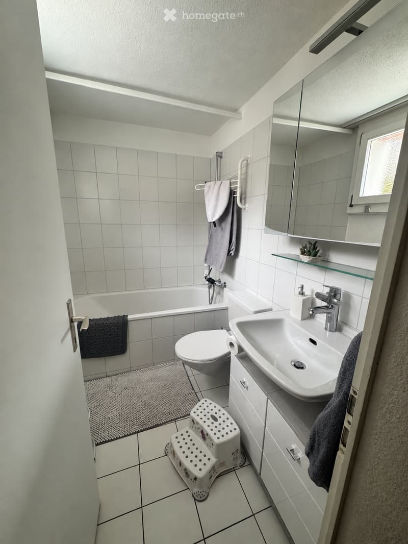 A small, well-lit bathroom with white tiles, a bathtub, toilet, sink, mirror, and a towel rack with towels. A small step stool is placed near the toilet.