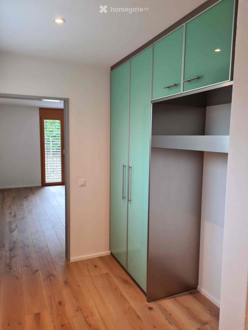 Empty room with light wooden flooring, white walls, and a set of modern green and grey cabinets with metallic handles. A doorway leads to another room with a glass-paneled door.