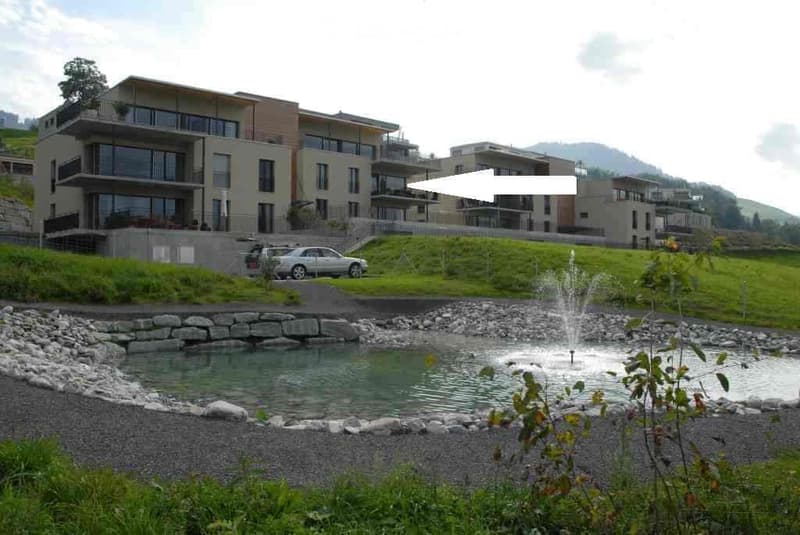 Modern multi-story residential building with balconies and large windows, situated on a hillside with a small pond and fountain in the foreground.