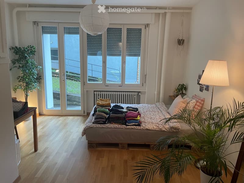 Bright bedroom with wooden flooring, large window with blinds, bed with folded clothes, floor lamp, and potted plants.