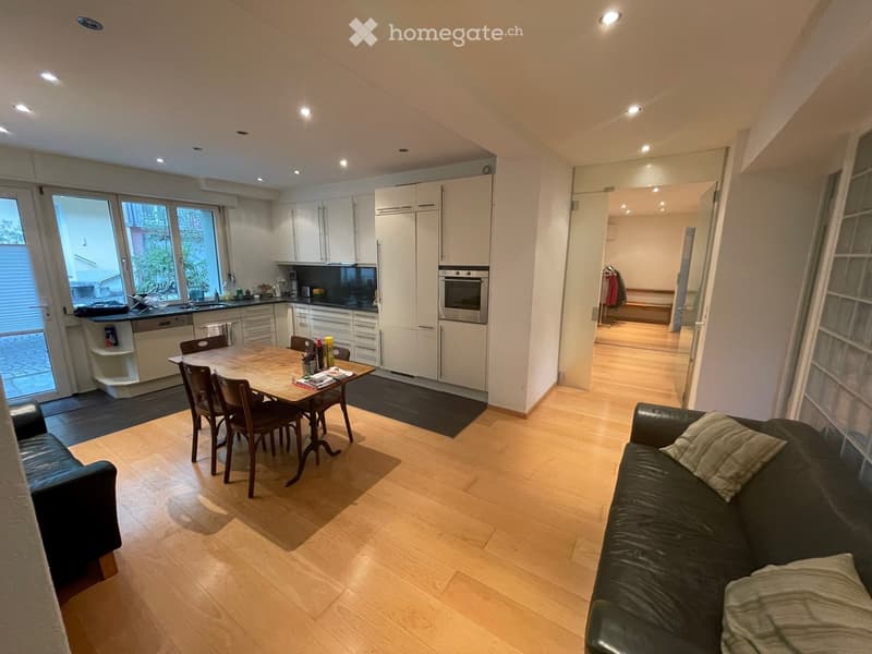 Open-plan living and dining area with light wooden flooring, a black leather sofa, a wooden dining table with chairs, and a modern kitchen with white cabinets, an oven, and a dishwasher.