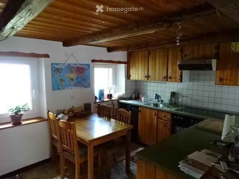 Cozy kitchen with wooden dining table and chairs, wooden cabinets, sink, and various appliances. Features a world map on the wall and a small plant on the windowsill.