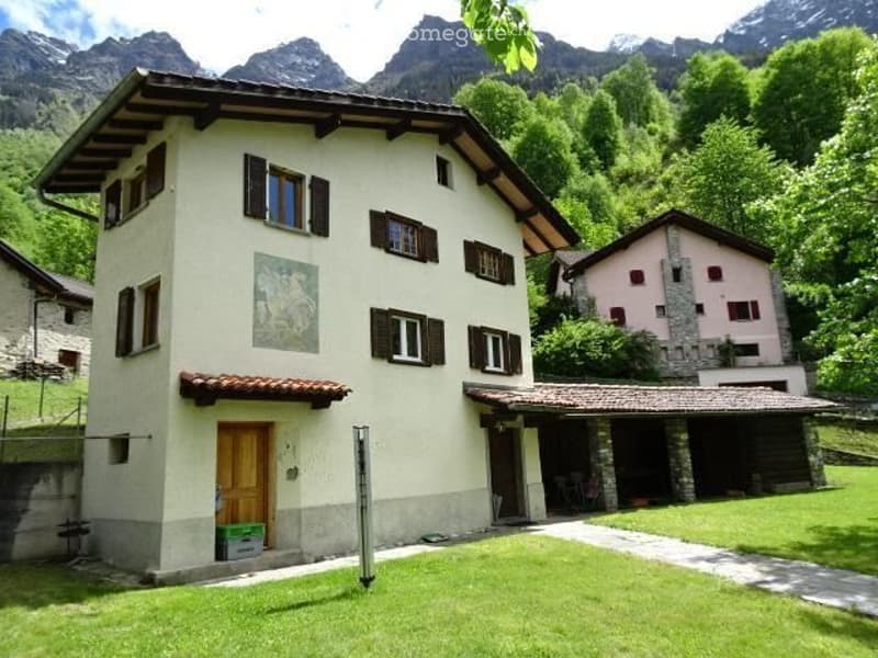 Two-story house with a cream-colored facade, wooden shutters, and a tiled roof, situated in a lush green area with mountain views in the background.