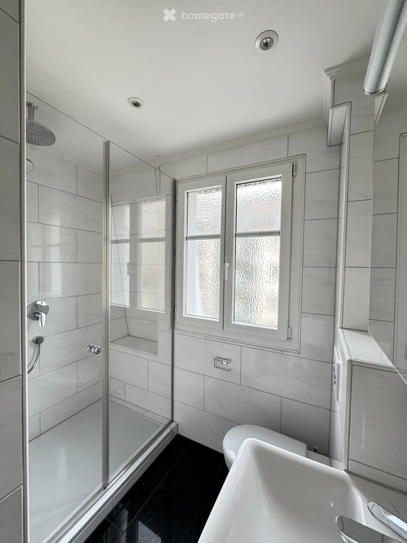 Modern bathroom with a white bathtub, toilet, glass-enclosed shower, and frosted glass window. Features sleek fixtures and dark tiled flooring.