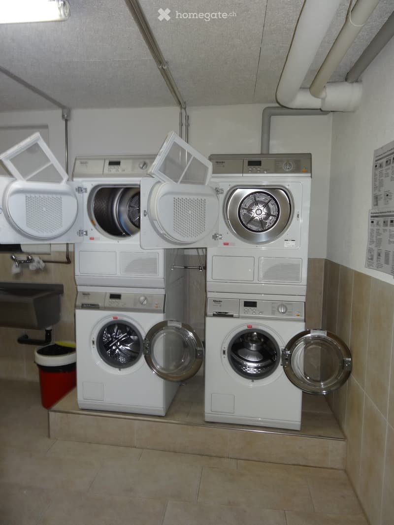 A small utility room with two stacked washing machines and dryers, beige tiled floor, white walls, and exposed pipes.