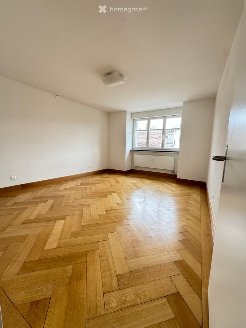Empty room with wooden herringbone flooring, white walls, and a window with a radiator below it.