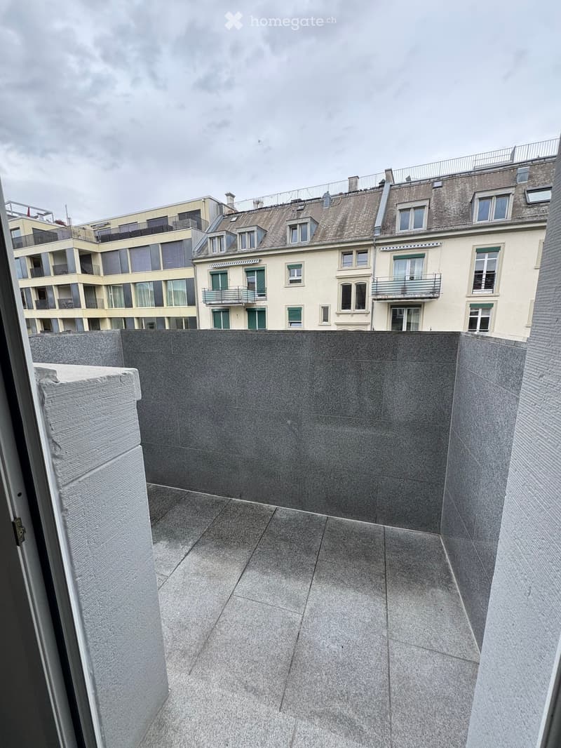 A modern rooftop terrace with grey stone tiles, low walls, and a view of surrounding buildings.