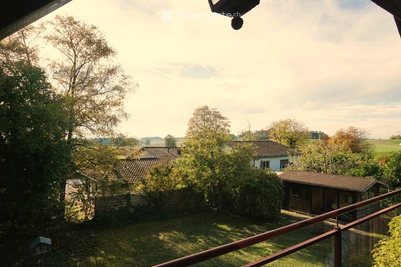 Balcony view overlooking a garden area with a small wooden shed, a house, and surrounding trees and fields.