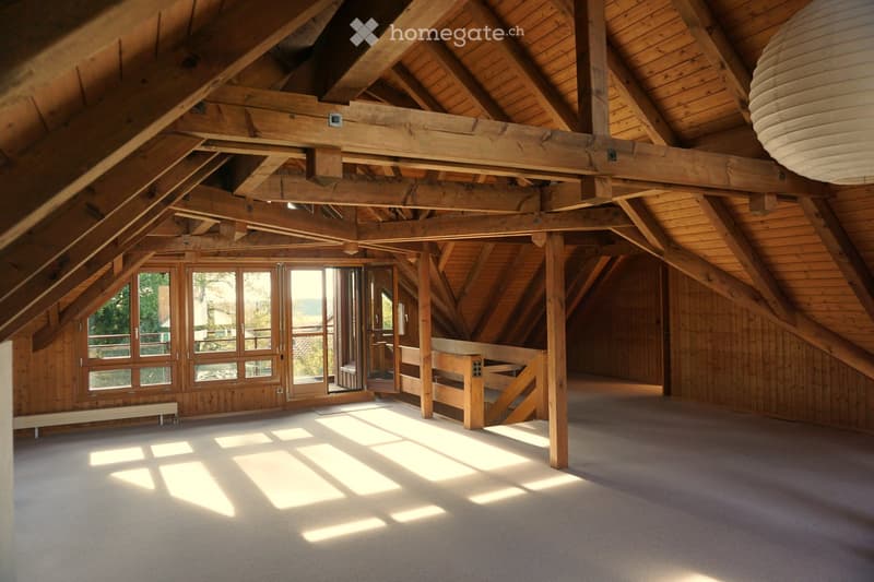 Spacious, open attic room with exposed wooden beams and large windows, featuring a wooden staircase leading to an upper level.