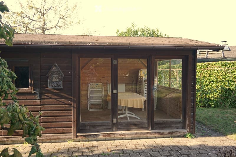 A wooden summerhouse with large glass doors, featuring a table and chairs inside, surrounded by greenery and a brick pathway.
