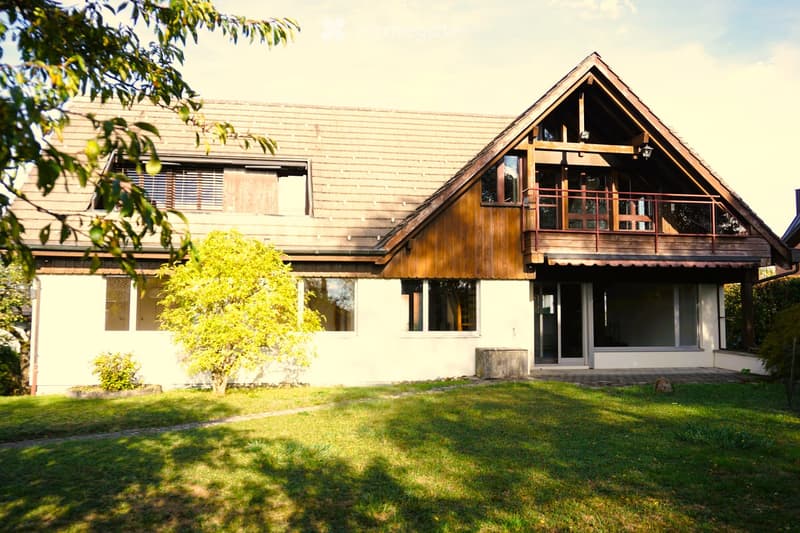 Two-story house with a wooden exterior, large windows, and a balcony on the upper level. The house features a well-maintained lawn and small garden area in front.