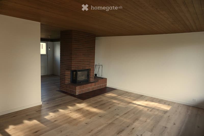 Empty room with wooden flooring, white walls, and a brick fireplace with a black insert. Sunlight streams in from a window on the left side.