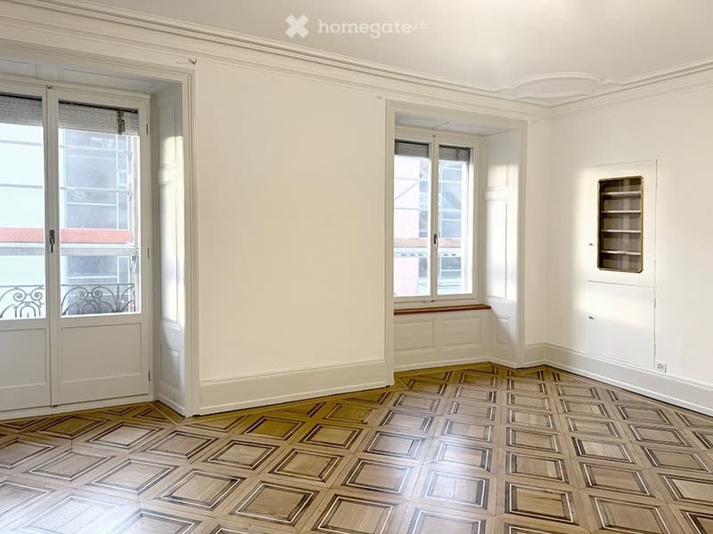 Empty room with white walls, ornate ceiling molding, and a patterned wooden floor. Features include a built-in window seat, a small built-in shelf, and large windows with blinds.
