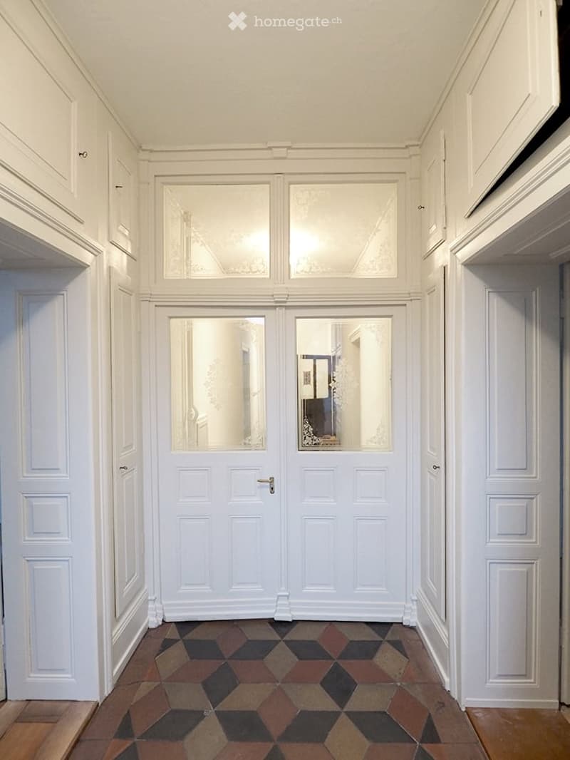 Elegant hallway with white paneled walls, geometric patterned tile floor, double doors with glass panels, and built-in cabinets.