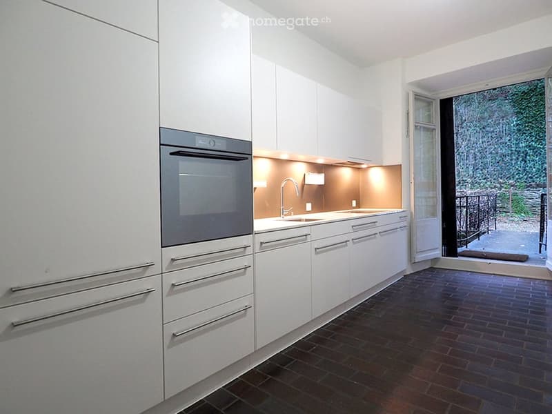 Modern kitchen with white cabinets, stainless steel oven, sink, and faucet, dark tiled floor, and a view of a garden through an open door.