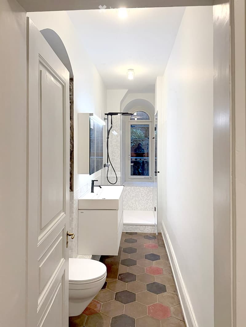 Modern bathroom with white walls, hexagonal tile floor, toilet, sink, mirror, and shower area with glass enclosure.