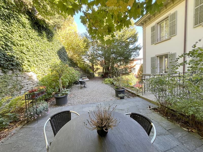 A serene garden area with a stone pathway, a small table with chairs, potted plants, and a view of a building with shutters.