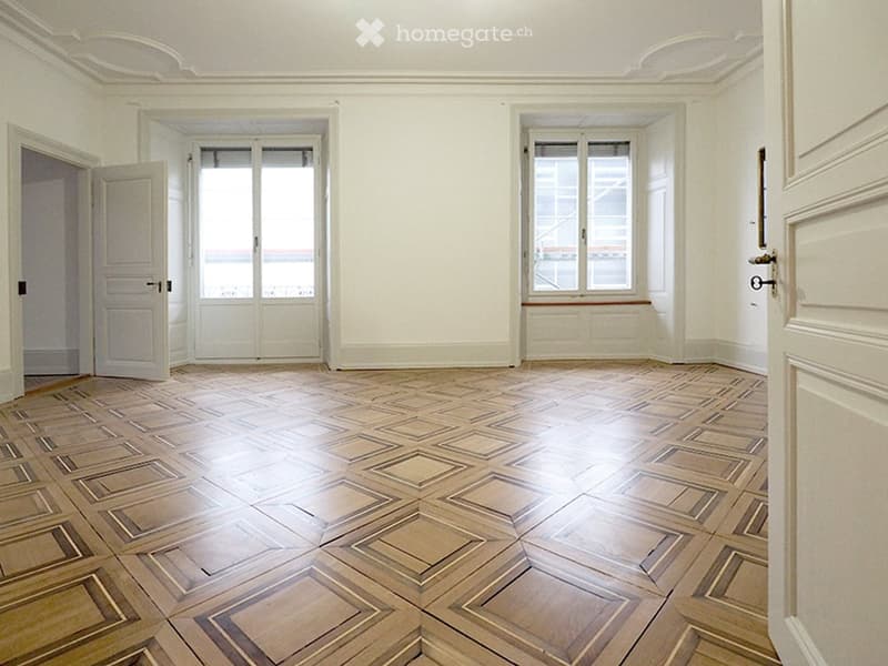 Empty room with wooden parquet flooring, white walls, and large windows with shutters. No furniture or equipment present.