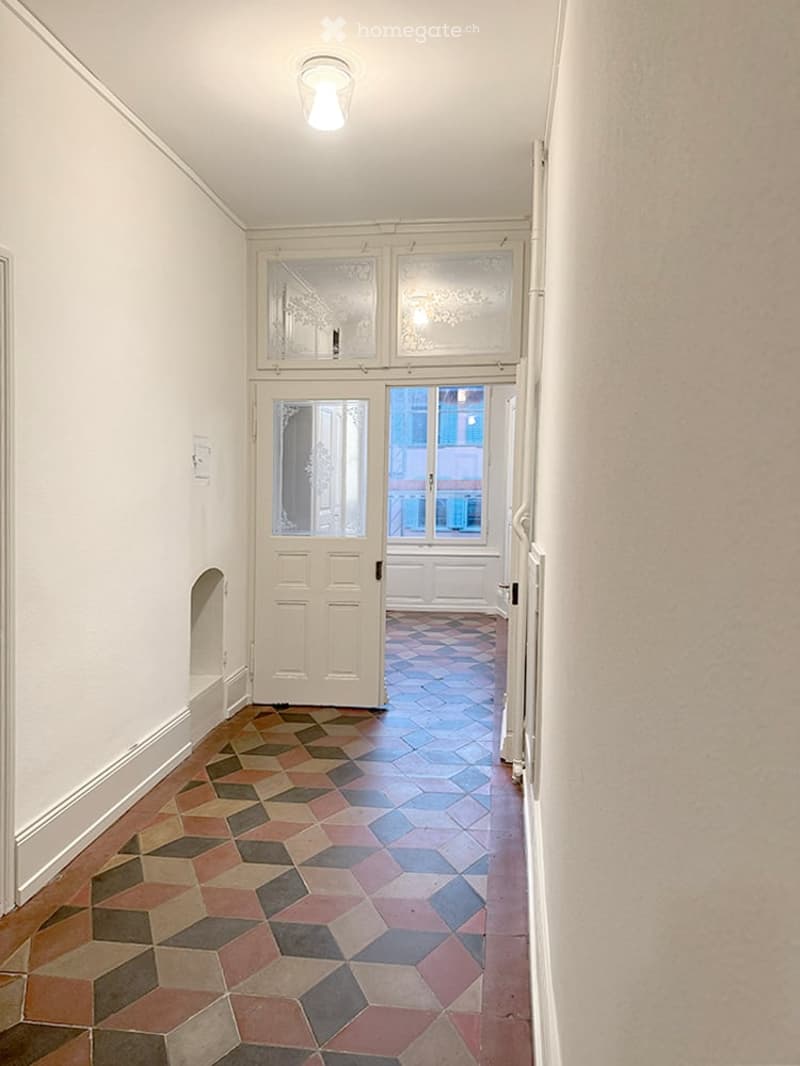 Empty hallway with patterned tile flooring, white walls, and a single ceiling light fixture. A door leads to another room with visible windows and a different tile pattern.