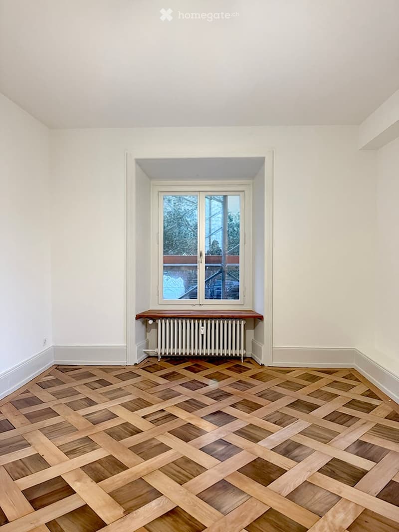Empty room with white walls, wooden herringbone floor, window with view, radiator, and small wooden bench.