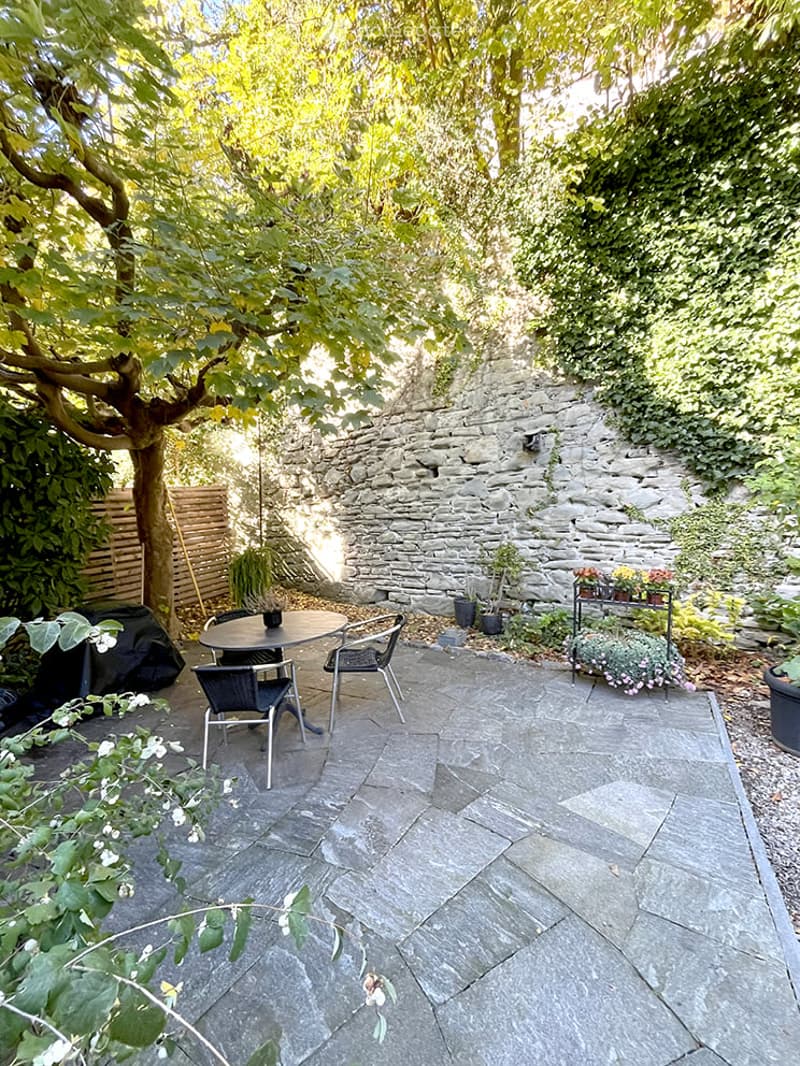 A small, enclosed garden patio with a stone floor, a round table with four chairs, potted plants, and a stone wall partially covered with ivy.