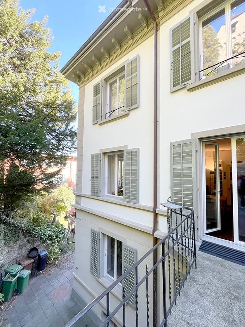 A multi-story residential building with white walls, multiple windows with shutters, and a small balcony accessible from the upper floor. The building is surrounded by greenery and has a paved patio area with trash bins.