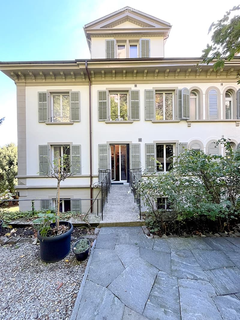 A two-story white house with classic architectural details, including shutters, a central entrance, and a small garden area with potted plants.