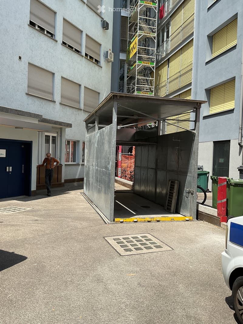 Multi-story building with a covered parking area, scaffolding on one side, and a man walking near the entrance.