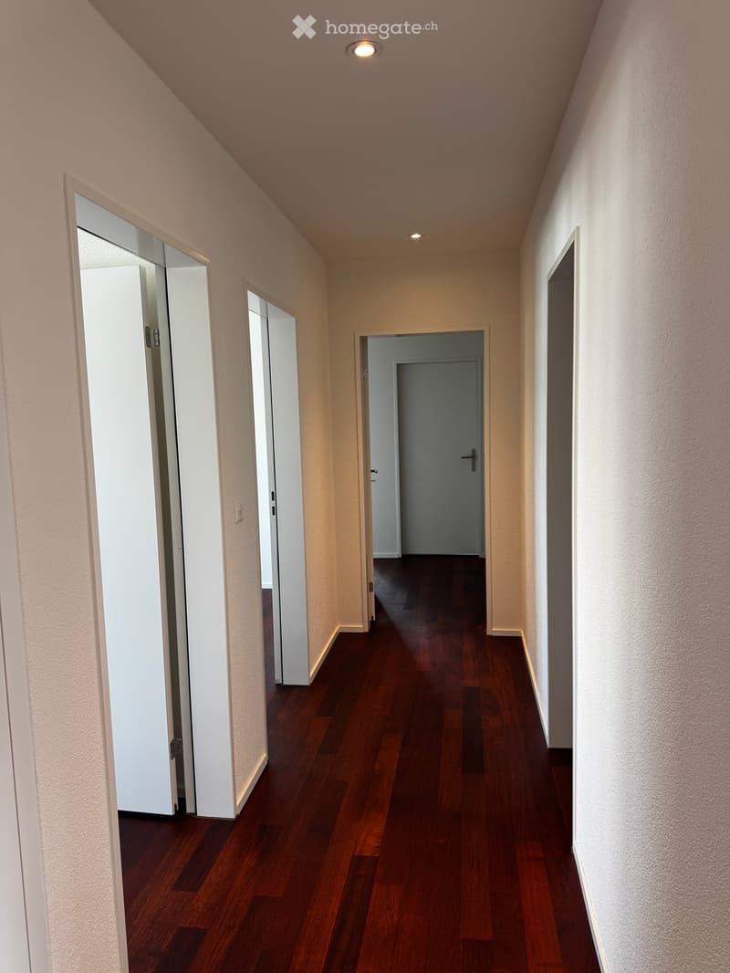 A narrow hallway with wooden flooring, white walls, and several doors leading to different rooms.
