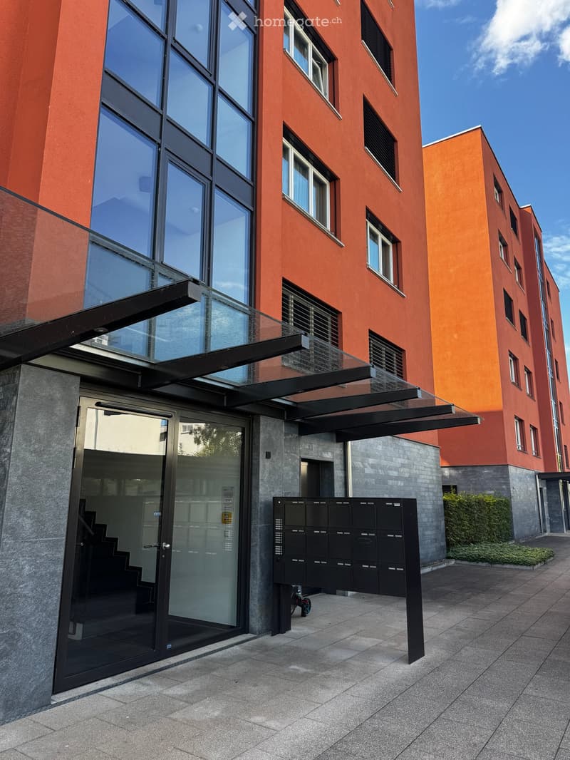Modern multi-story building with red brick facade, large windows, glass entrance, and covered walkway.