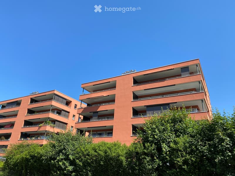 Modern multi-story apartment building with red brick facade, multiple balconies, and greenery in the foreground.