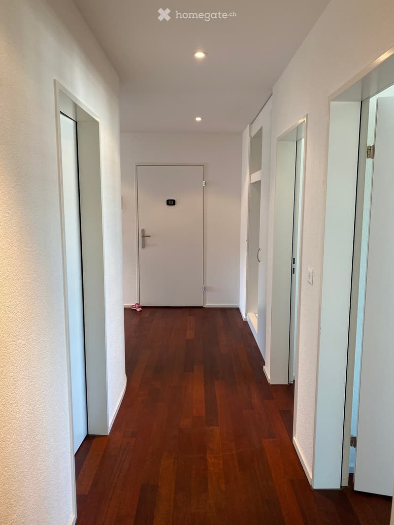 A clean, modern hallway with wooden flooring, white walls, and several doors, including a central closed door and mirrored glass doors on the right side.