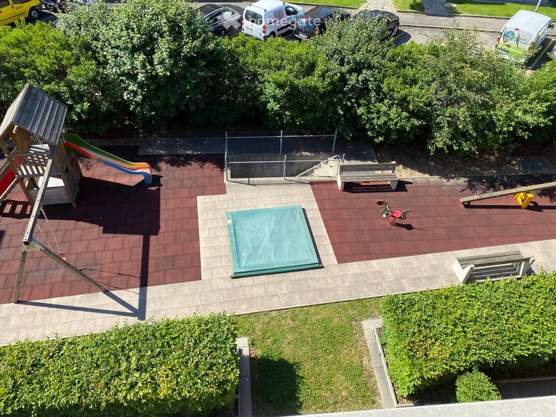 Aerial view of a playground area with a slide, seesaw, benches, and a covered area, surrounded by greenery and a parking lot with several cars.