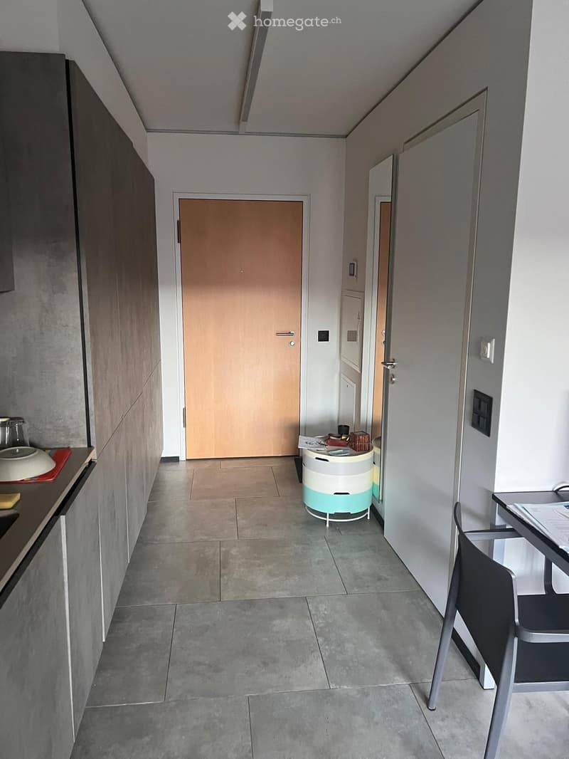 A narrow hallway with tiled flooring, grey walls, and several doors leading to different rooms. There is a small round table with a container on it, and a chair near the end of the hallway.