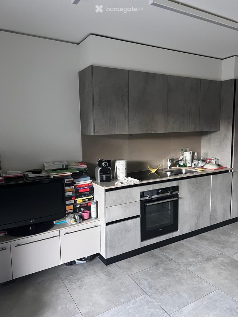 Modern kitchen with grey cabinets, stainless steel appliances, and a tiled floor. Includes an oven, sink, coffee maker, and a TV on a stand with books and cups.