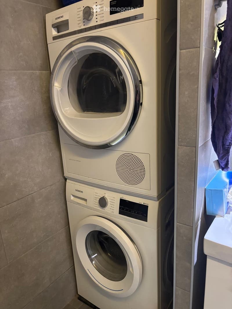 A compact bathroom with a stacked washing machine and dryer unit, beige tiled walls, and a small shelf with a folded towel and a blue box.