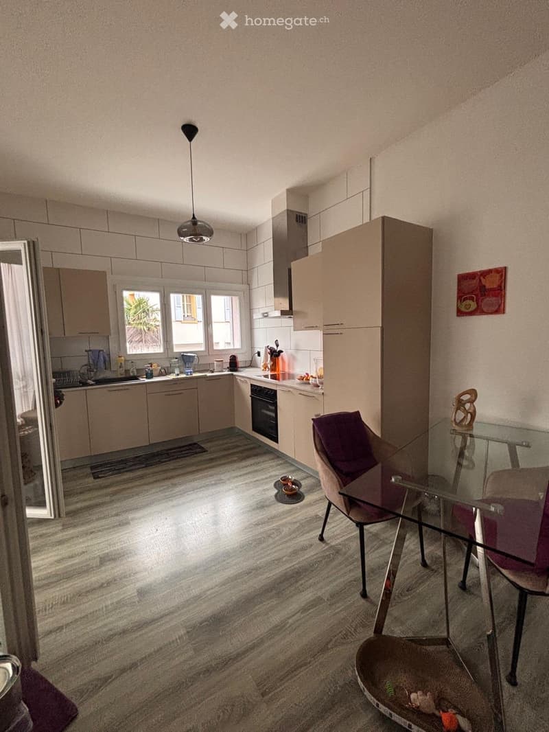 A modern kitchen with light-colored cabinets, a glass dining table with chairs, and various appliances including a sink, oven, and refrigerator. The floor is made of polished wood, and there is a pendant light fixture above the dining area.