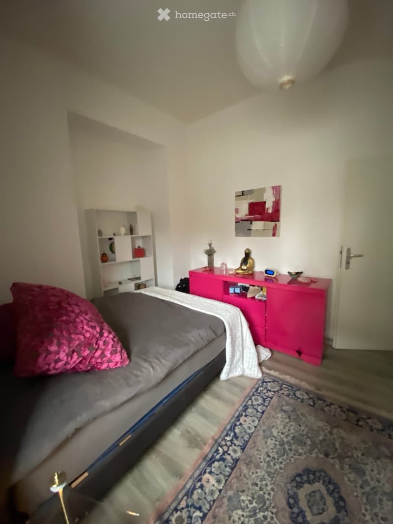 A modern bedroom with a grey bed, pink nightstand, white shelf, and a patterned rug.