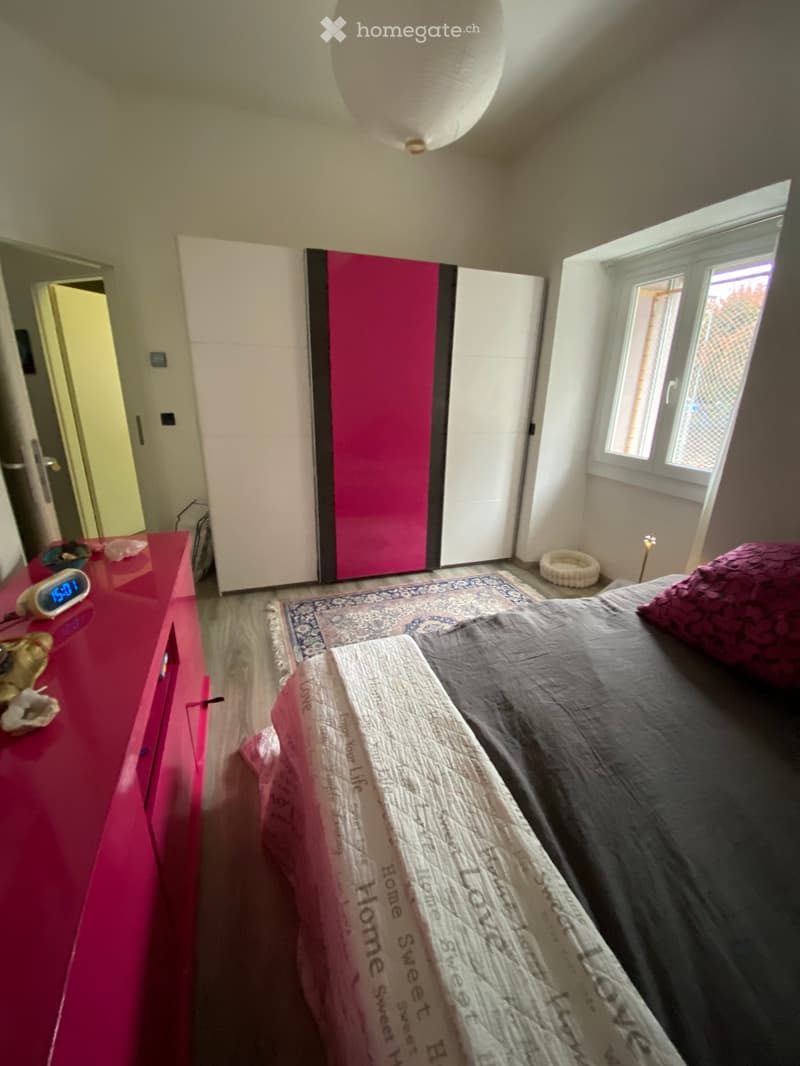 A modern bedroom with a large bed, pink nightstand, white wardrobe with pink accents, and a pet bed in the corner.