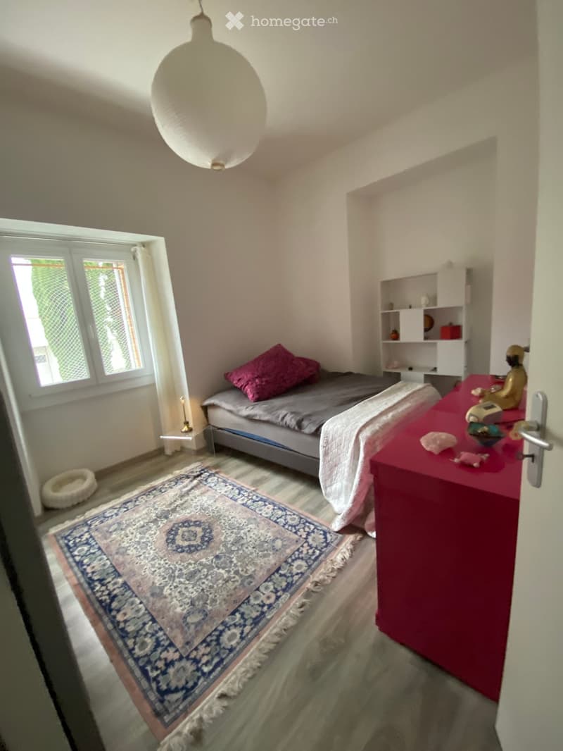 Cozy bedroom with a bed, colorful pillow, patterned rug, small table with decorative items, and a shelf with books and decorative objects.