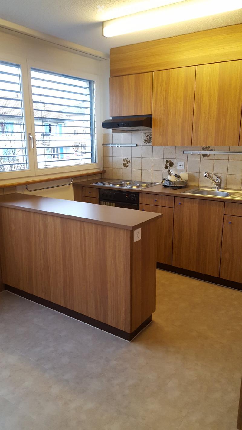 Modern kitchen with wooden cabinets, a central island, and stainless steel appliances including a sink, oven, and range hood. The space features tiled backsplash and large windows.