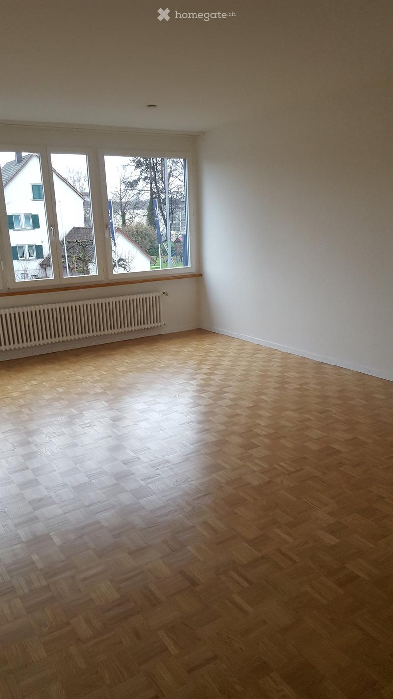 Empty room with wooden herringbone flooring, white walls, large windows, and a radiator below the windows.