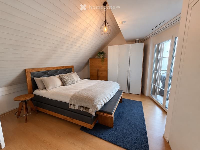 Cozy attic bedroom with wooden bed, bench seating, white wardrobe, and large windows offering natural light.