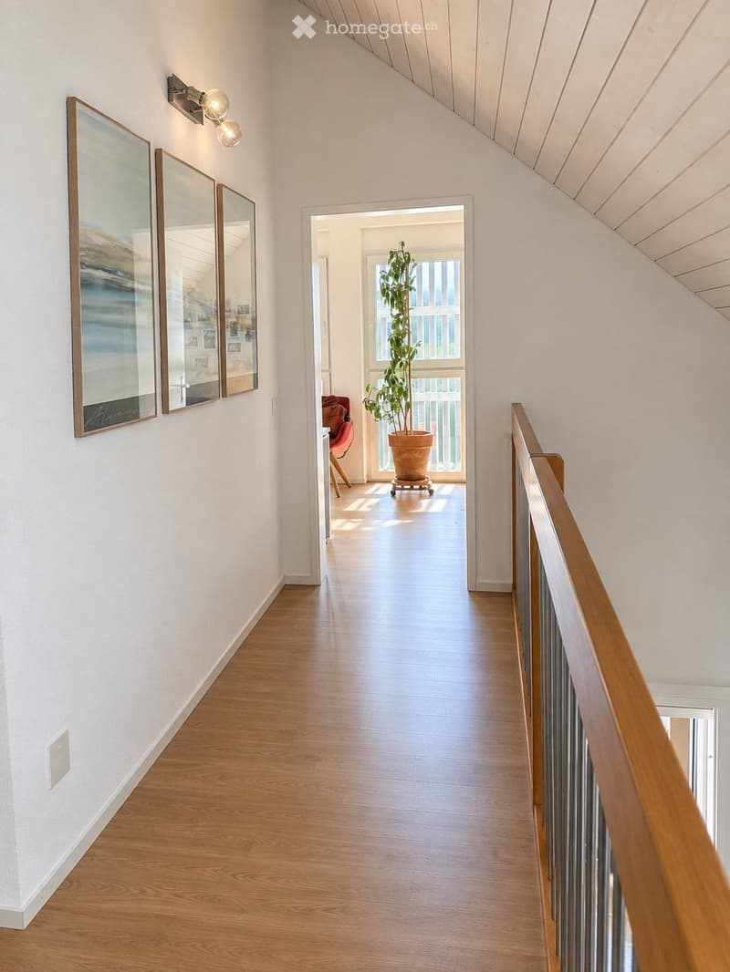 Bright hallway with wooden flooring, white walls, and a wooden railing on the right side. Three framed mirrors are mounted on the left wall, and a wall-mounted light fixture is visible. The hallway leads to an open area with a potted plant and a red chair