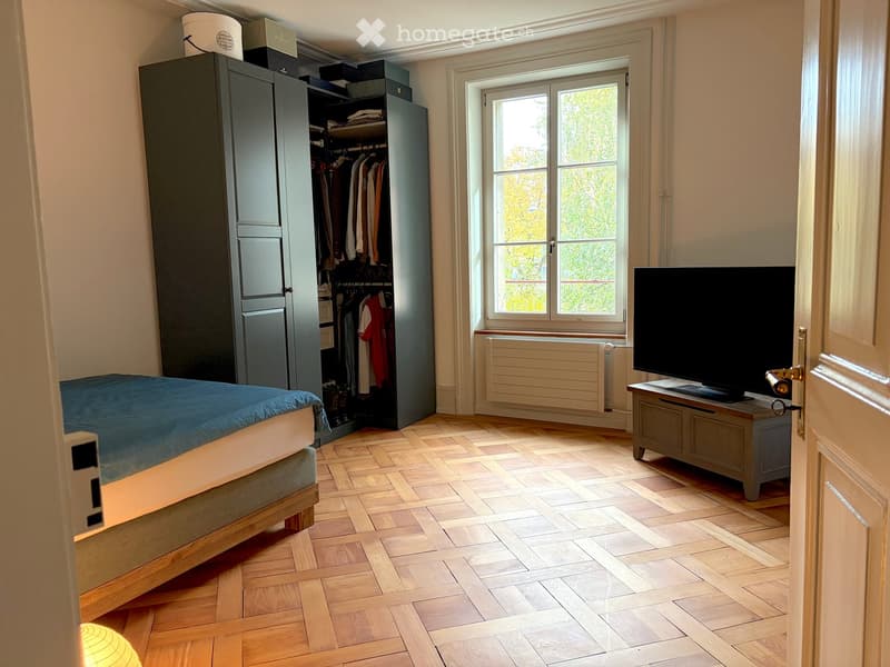 Spacious bedroom with wooden herringbone flooring, large window with view, bed with blue duvet, wardrobe with hanging clothes, TV on a stand, and a small table.