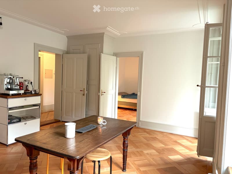 A well-lit kitchen area with a wooden table, a stool, a coffee machine, an oven, and a refrigerator. The room has light-colored walls and wooden flooring, with an open doorway leading to another room.