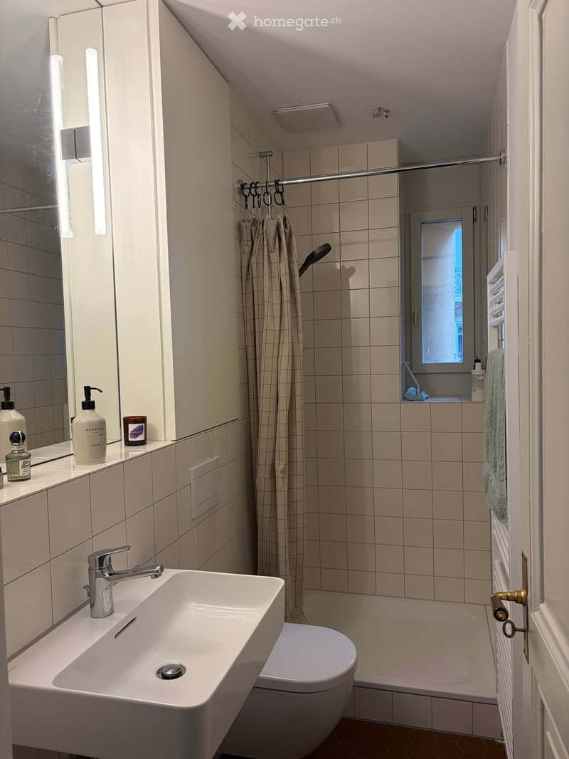Modern bathroom with a white sink, chrome faucet, toilet, bathtub, and shower curtain. A mirror with toiletries and a candle sits above the sink. A towel hangs on the radiator.