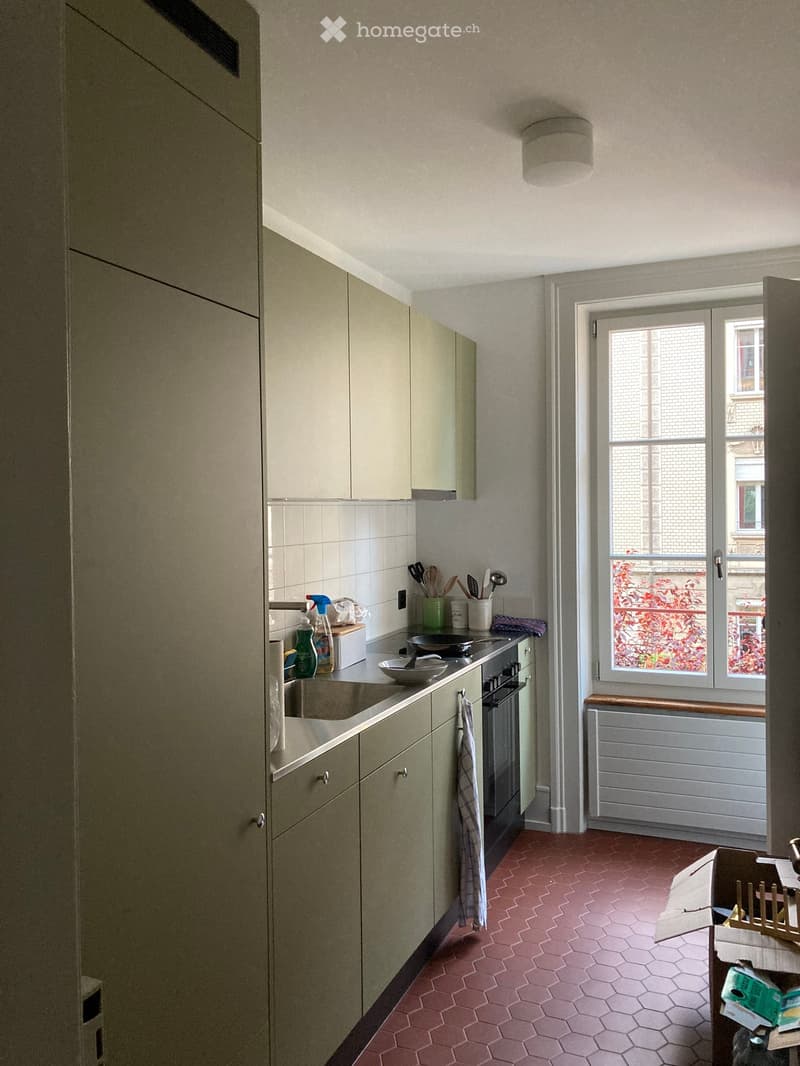 The kitchen features light-colored cabinets, a stainless steel sink, and a stove with a towel hanging on the oven handle. The floor is tiled in a pink hexagonal pattern, and a window with a view of a brick building is visible.