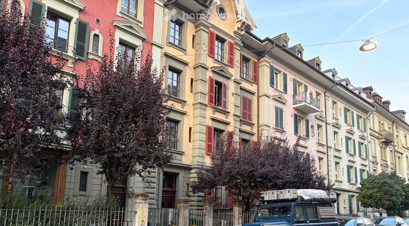 A row of multi-story residential buildings with colorful facades, green shutters, and decorative architectural details, featuring trees and parked vehicles in front.